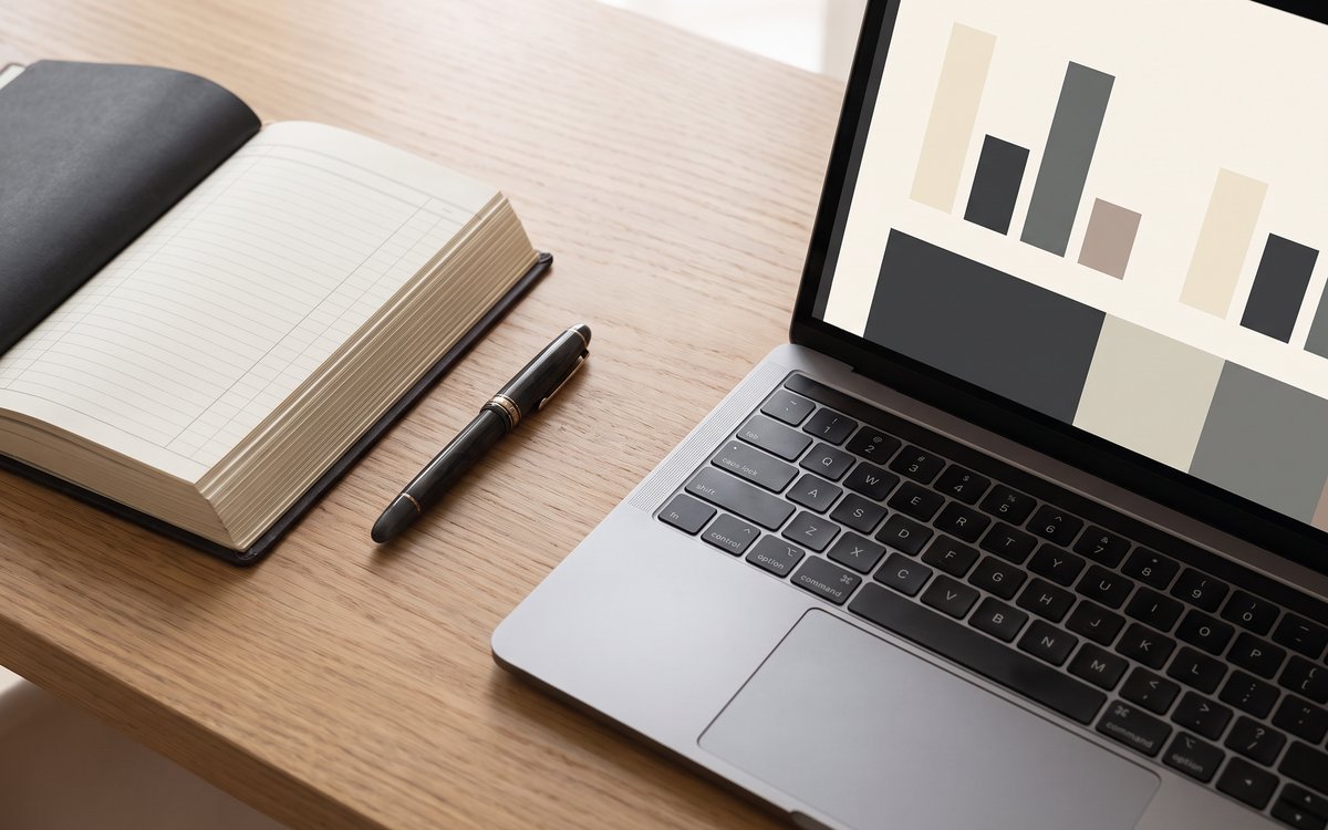 An organized desk with a bound ledger notebook and pen beside a laptop showing simple charts, in soft natural light.